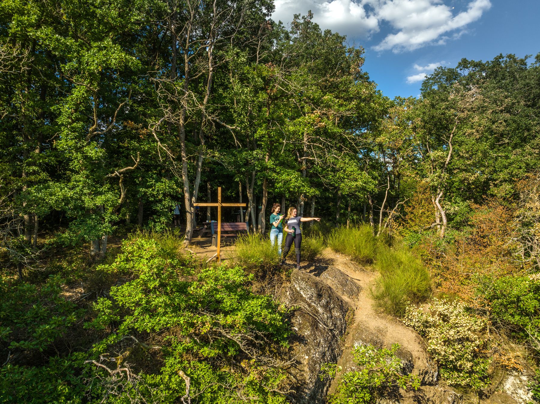 Zwei Personen stehen auf einem felsigen Weg, der von dichten grünen Bäumen umgeben ist, wobei eine Person nach vorne zeigt. In der Nähe stehen ein großes Holzkreuz und eine Bank, die teilweise im Schatten der Bäume zu sehen ist. Der Himmel ist teilweise bewölkt.